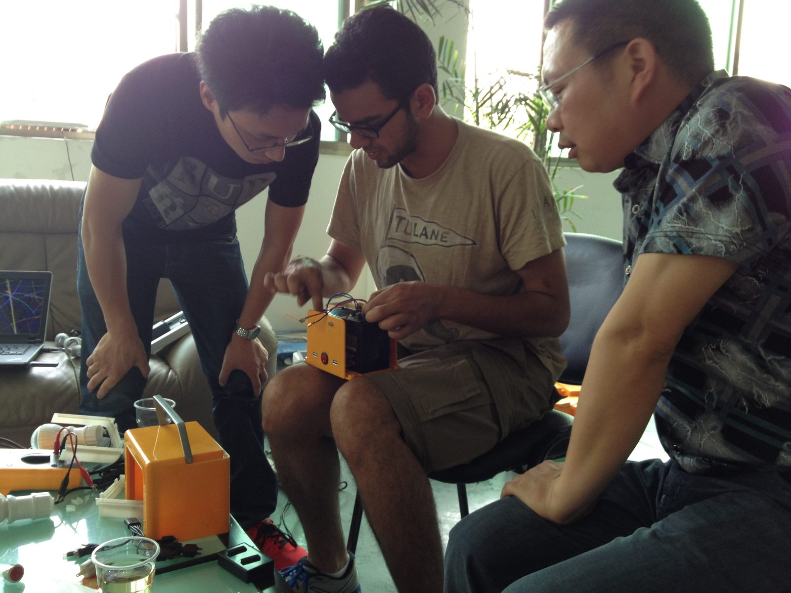 Three people working through a BrightBox prototype on a couch in a Chinese manufacturing office, with circuit boards and parts on the table