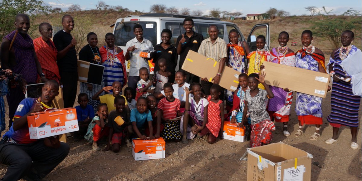 Group of villagers, mostly women in colorful headscarves, gathered around a young man holding an orange BrightBox while a woman in red explains the product