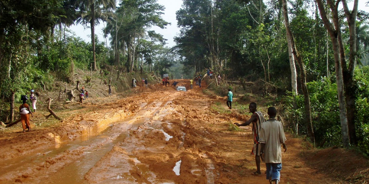 Rural road in Liberia, washed out and muddy after rain, with people walking and a vehicle stuck in deep ruts