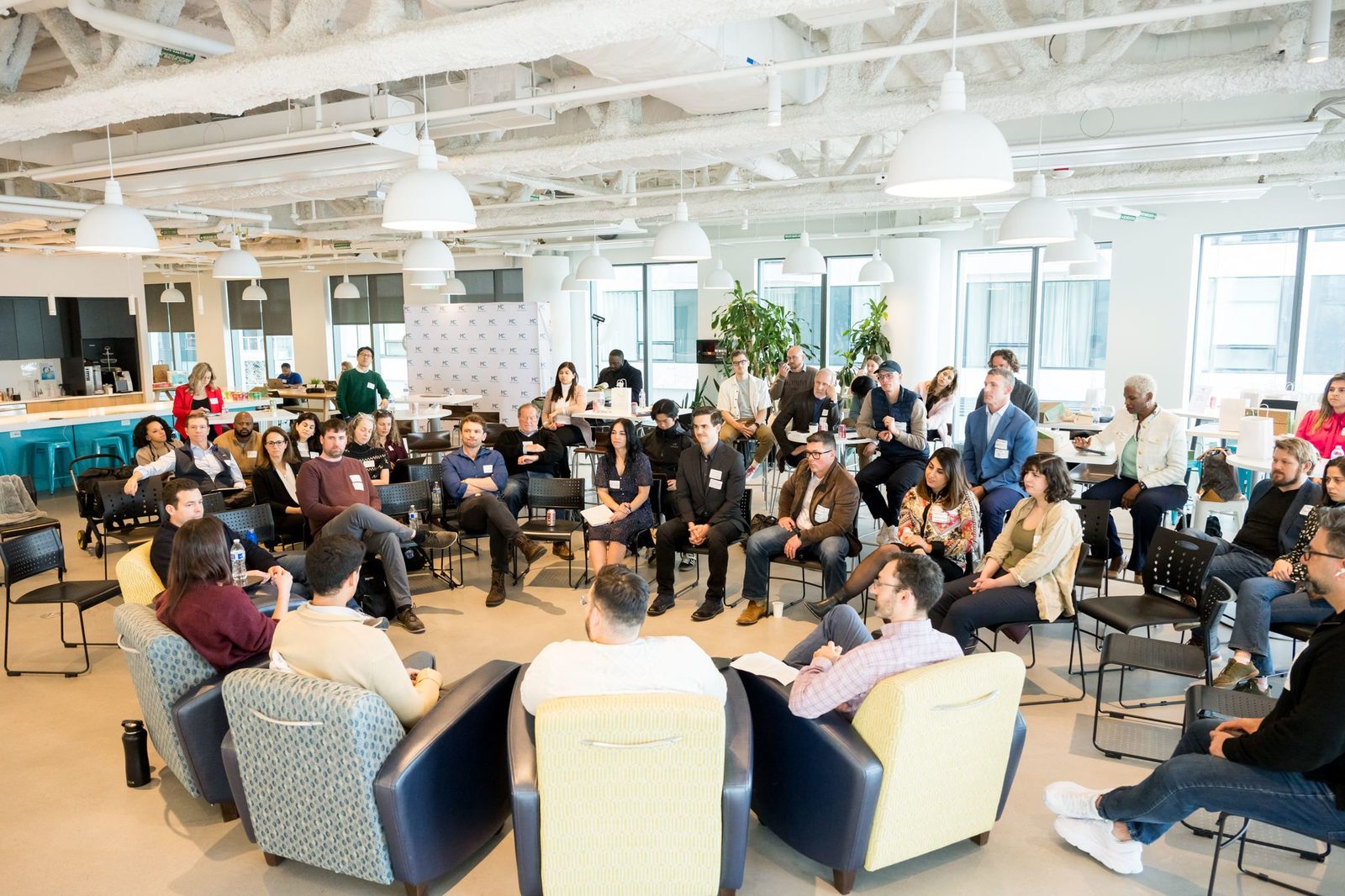 MassChallenge cohort gathered in a large open accelerator space, founders seated in a U-shape facing presenters in the center